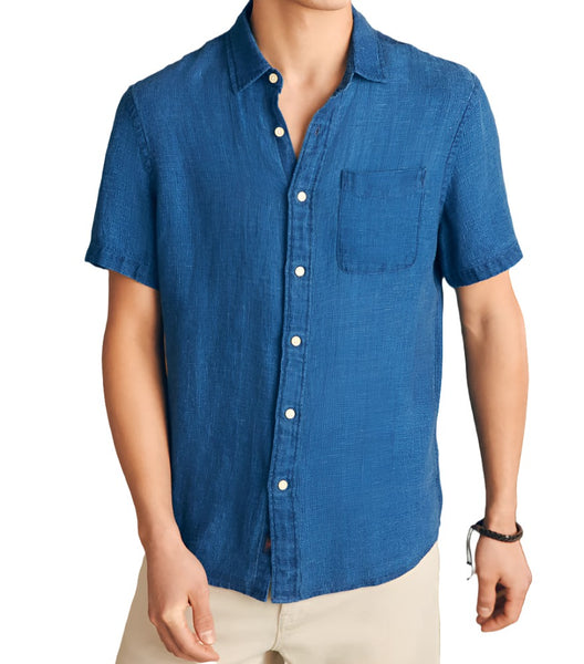 A person is wearing a Faherty Short-Sleeve Linen Basketweave Shirt, a blue short-sleeved button-up with a pocket on the left chest and light-colored pants. They have a black bracelet on their right wrist and are standing against a plain background.