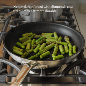 The Hestan Thomas Keller Commercial Clad Stainless Steel TITUM® Nonstick Sauté Pan, 12.5", features a ProCore™ aluminum core and diamond-titanium reinforcement for 22x more durability, shown here with asparagus on a gas burner.