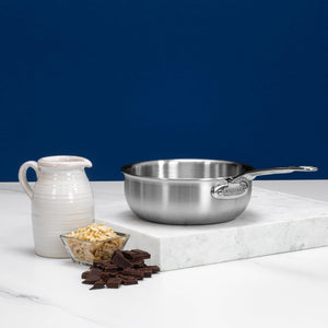The Hestan Thomas Keller Commercial Clad Stainless Steel Saucier, 2qt, sits on a marble slab beside a white ceramic pitcher, a glass bowl of slivered almonds, and dark chocolate pieces against a blue background.