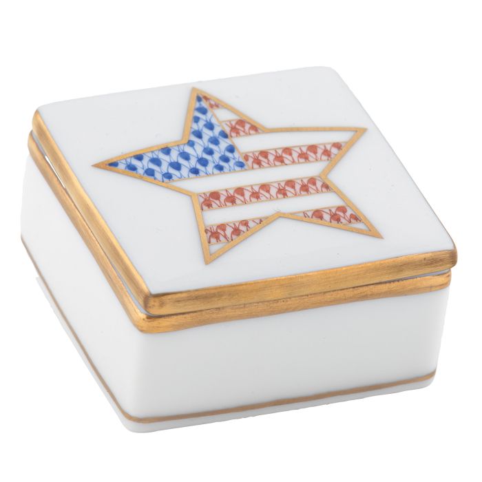 The Herend Patriotic Box is a small, square white porcelain collectible with gold trim and a hand-painted lid featuring a star in blue, red, and white stripes reminiscent of the American flag—a charming accent.