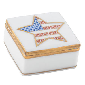 The Herend Patriotic Box is a small, square white porcelain collectible with gold trim and a hand-painted lid featuring a star in blue, red, and white stripes reminiscent of the American flag—a charming accent.