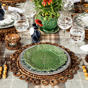 The Juliska Rustic Ring Placemat, paired with green patterned plates, glassware, bamboo-handled cutlery, a potted plant centerpiece, and a checked tablecloth, creates an inviting and textured table setting that beautifully enhances your rustic decor.