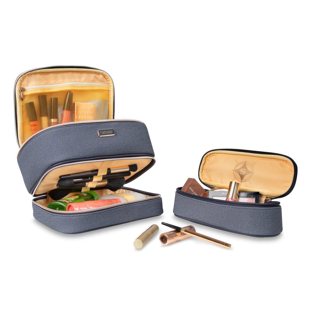 Two makeup bags from the Stephanie Johnson Denim Travel Collection, shown open like sleek cosmetic cases with visible beige lining, each holding assorted makeup items and toiletries.