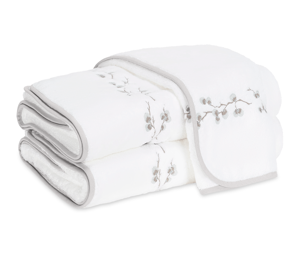 Two folded white towels from the Matouk Jasmine Giza Bath Collection in Cloud, featuring light gray edging and a subtle branch and blossom embroidery, crafted from plush Egyptian cotton terry for a luxurious bathroom experience.