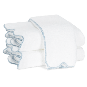 Two folded white towels with light blue trim from the Matouk Jasper Bath Collection, featuring a subtle scalloped silhouette, are stacked on a plain white background.