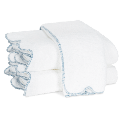 Two folded white towels with light blue trim from the Matouk Jasper Bath Collection, featuring a subtle scalloped silhouette, are stacked on a plain white background.