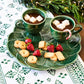Two green cups of hot chocolate with marshmallows, served with strawberries, cookies, and a mini candy cane, are set on Juliska's Le Panier Mystic Knot Basil Dinnerware—a festive choice for your holiday table.