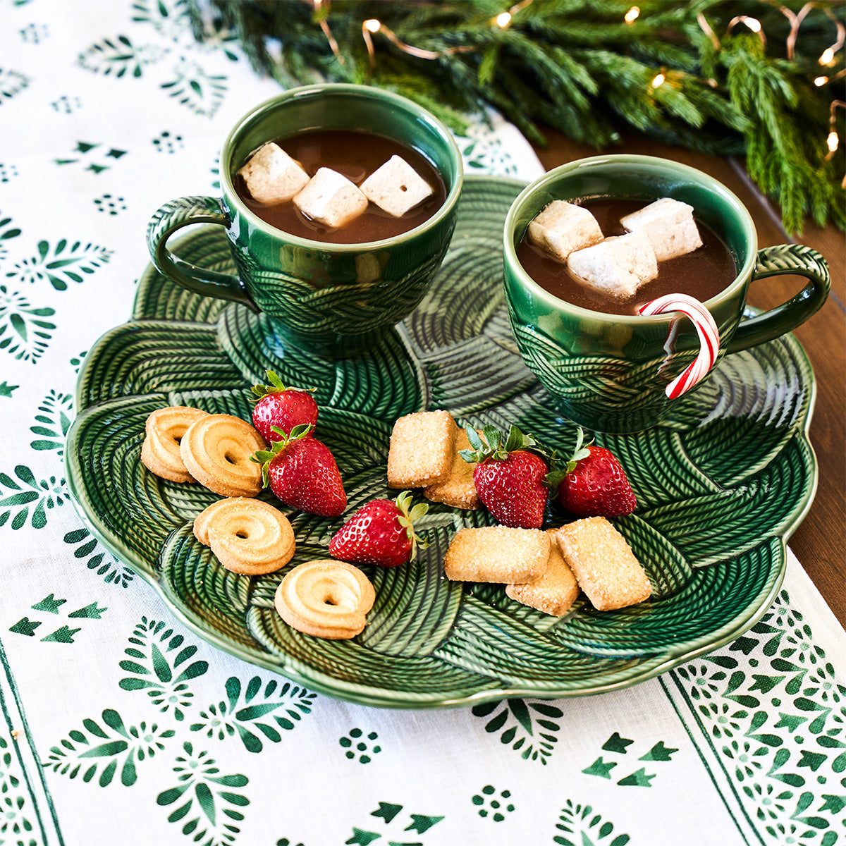 Two green cups of hot chocolate with marshmallows, served with strawberries, cookies, and a mini candy cane, are set on Juliska's Le Panier Mystic Knot Basil Dinnerware—a festive choice for your holiday table.