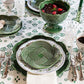 A table set with Juliska’s Le Panier Mystic Knot Basil Dinnerware and Serveware Collection, featuring patterned green and white plates and bowls, glasses, silver cutlery, and a green leaf tablecloth with ceramic bowls holding food and drinks.