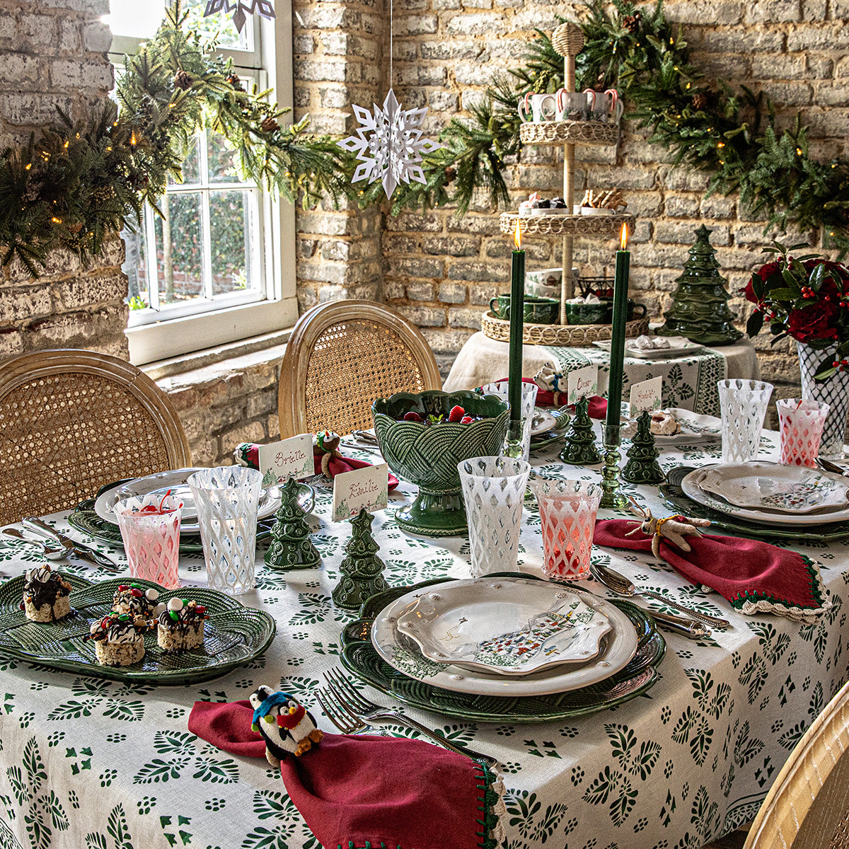 A holiday table is set with Juliska’s Le Panier Mystic Knot Basil Dinnerware and Serveware Collection, Christmas tree decorations, candles, and festive garland by a sunlit window.