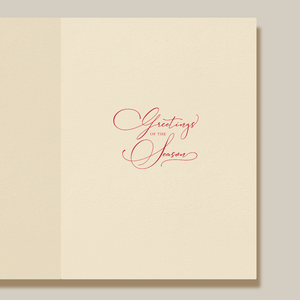 The Christmas Morning Wreath Card Set by Crane features an open cream-colored card with "Greetings of the Season" in red cursive at the center, framed by a delicate wreath design.