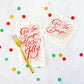 Two cream-colored Hester & Cook ’Tis the Season Napkin Collection napkins with red and gold text, accented by a gold fork and colorful confetti.