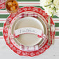 The Classic Red Banner Table Accent by Hester & Cook adds festive charm to your Christmas table, pairing beautifully with a red and white charger, beige plate, striped napkin, cutlery, and a drink for a complete holiday place setting.