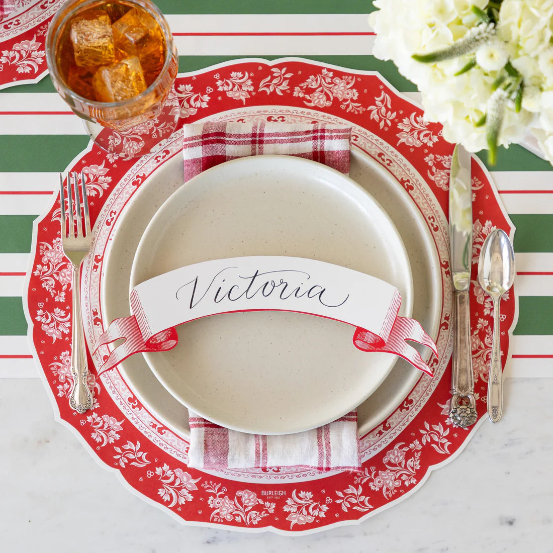 The Classic Red Banner Table Accent by Hester & Cook adds festive charm to your Christmas table, pairing beautifully with a red and white charger, beige plate, striped napkin, cutlery, and a drink for a complete holiday place setting.