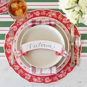 The Classic Red Banner Table Accent by Hester & Cook adds festive charm to your Christmas table, pairing beautifully with a red and white charger, beige plate, striped napkin, cutlery, and a drink for a complete holiday place setting.