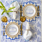 A formal table setting for two features Hester & Cook's Die-cut Bamboo Placemat Collection in blue and white, gold-edged plates, gold flatware, amber glasses, and white napkins with blue trim beside a vase of green flowers for festive decor.