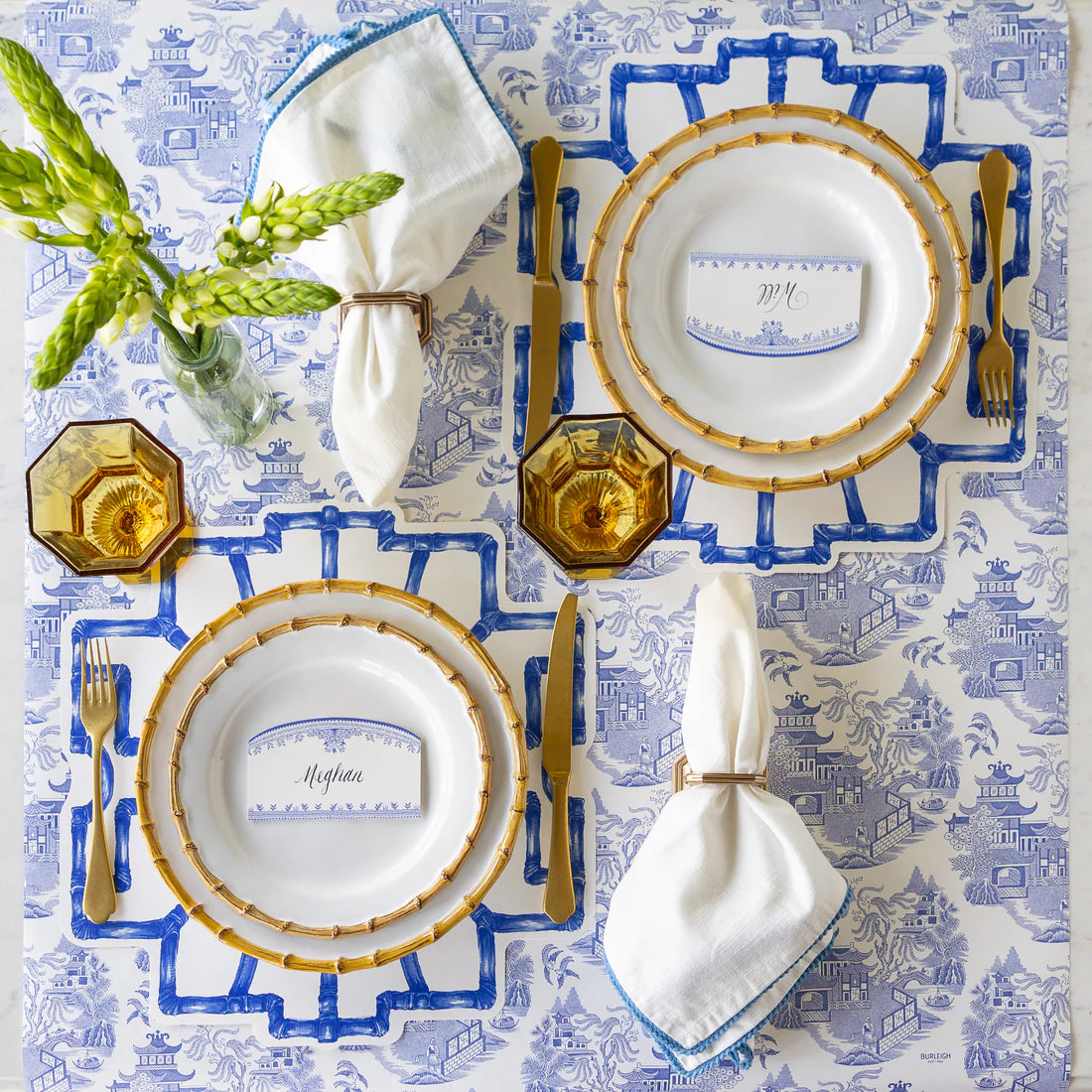 A formal table setting for two features Hester & Cook's Die-cut Bamboo Placemat Collection in blue and white, gold-edged plates, gold flatware, amber glasses, and white napkins with blue trim beside a vase of green flowers for festive decor.