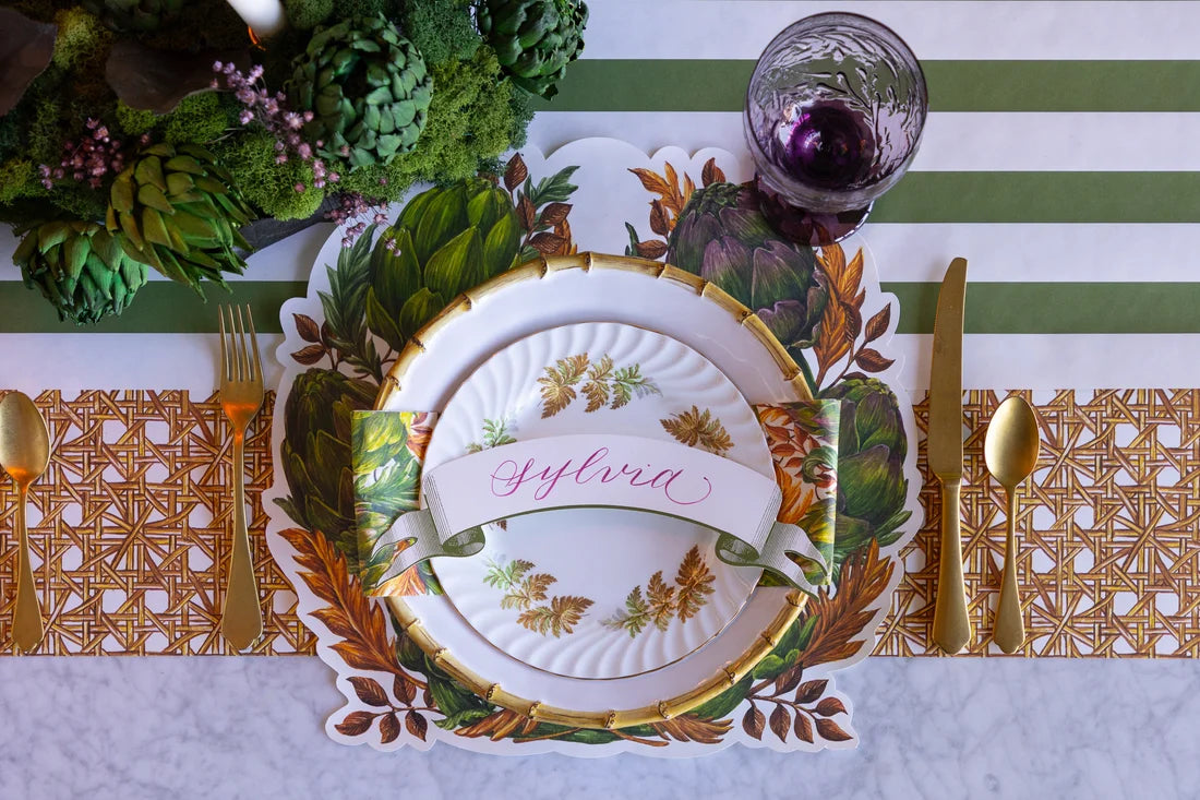 The Autumnal Artichoke decorative plate by Hester & Cook is set with gold utensils and a glass filled with purple liquid; warm fall colors enhance the scene, while a name card labeled “Sylvia” sits atop the plate.