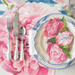 A table set with a fork, knife, and spoon beside a plate with blue trim and a "congrats" napkin, all atop the Hester & Cook Peony Collection placemat—ideal for a bridal brunch or garden party.
