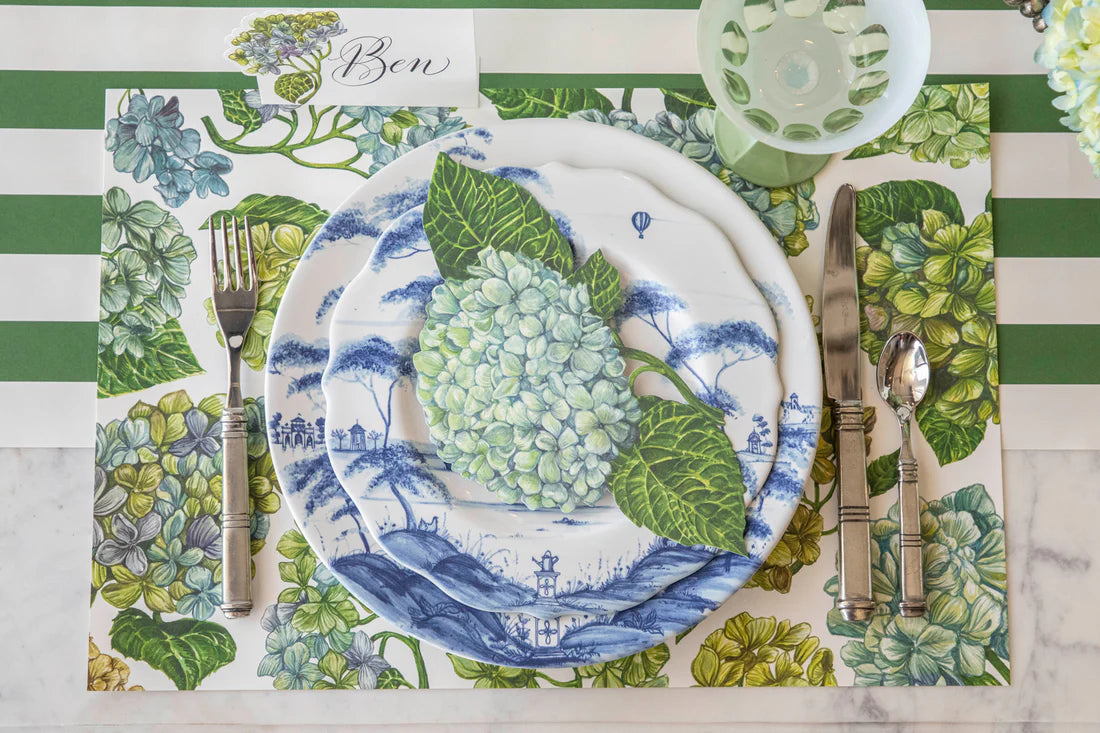 A table set with blue and white plates, silver cutlery, a green glass, and a floral placemat from Hester & Cook’s Hydrangea Collection features a hydrangea accent and a place card labeled "Ben" for an elegant setting.