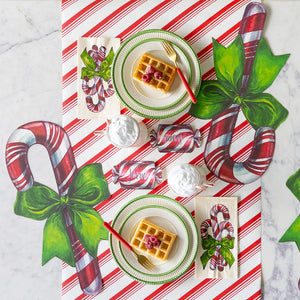 A festive tablescape featuring the Candy Cane Collection, with two plates of raspberry-topped waffles, peppermint-patterned placemats and napkins, and small decorative pillows labeled “Merry” and “Susan.”