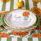 A place setting from the Marigold Collection by Hester & Cook showcases elegant white plates with bamboo-inspired rims, a fresh green napkin, and a “Luisa” name card adorned with a marigold illustration, all artfully set on a floral-patterned tablecloth—an ideal choice for your Thanksgiving gathering or any autumn-themed decor.