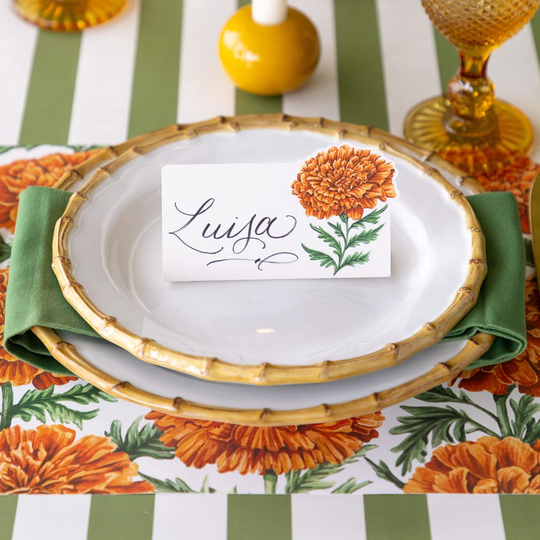 A place setting from the Marigold Collection by Hester & Cook showcases elegant white plates with bamboo-inspired rims, a fresh green napkin, and a “Luisa” name card adorned with a marigold illustration, all artfully set on a floral-patterned tablecloth—an ideal choice for your Thanksgiving gathering or any autumn-themed decor.