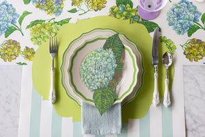 A table setting features a green placemat, scalloped-edge plate, Hester & Cook Hydrangea Table Accent place card, silver cutlery, and a folded blue napkin on a floral-striped tablecloth for charming decor.