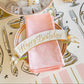 A pink napkin on a pink-rimmed plate with Hester & Cook's Happy Birthday Banner Table Accent, surrounded by festive decorations, illustrated utensils, and a bowl of ice cubes.