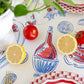 Tabletop styled with the Hester & Cook Seaside Party Runner featuring red and blue seafood motifs, sliced lemons, whole tomatoes, plate edges, and a white planter with greenery.