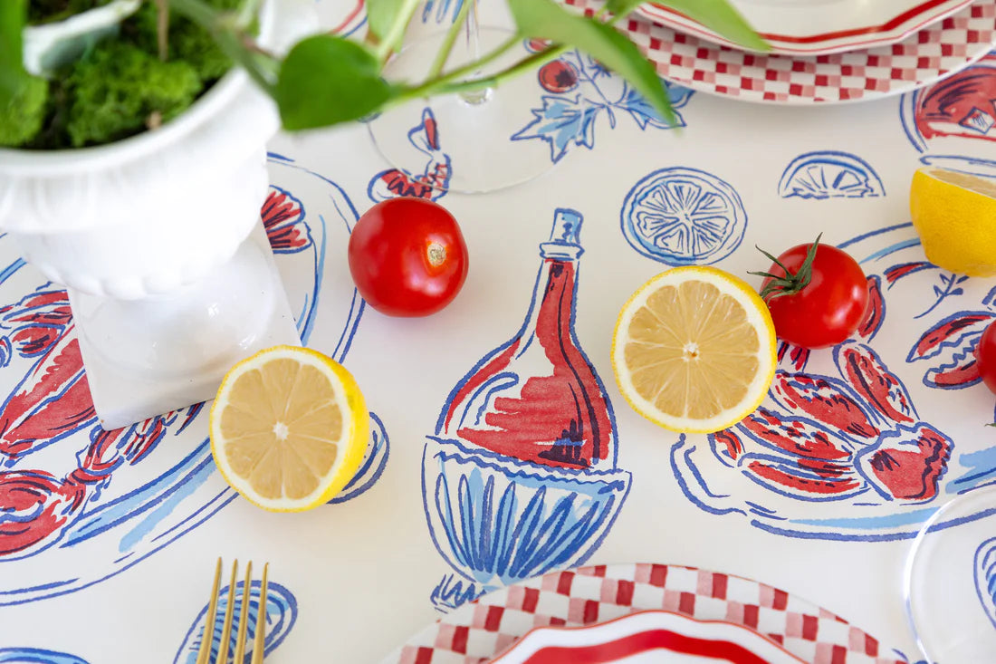 Tabletop styled with the Hester & Cook Seaside Party Runner featuring red and blue seafood motifs, sliced lemons, whole tomatoes, plate edges, and a white planter with greenery.