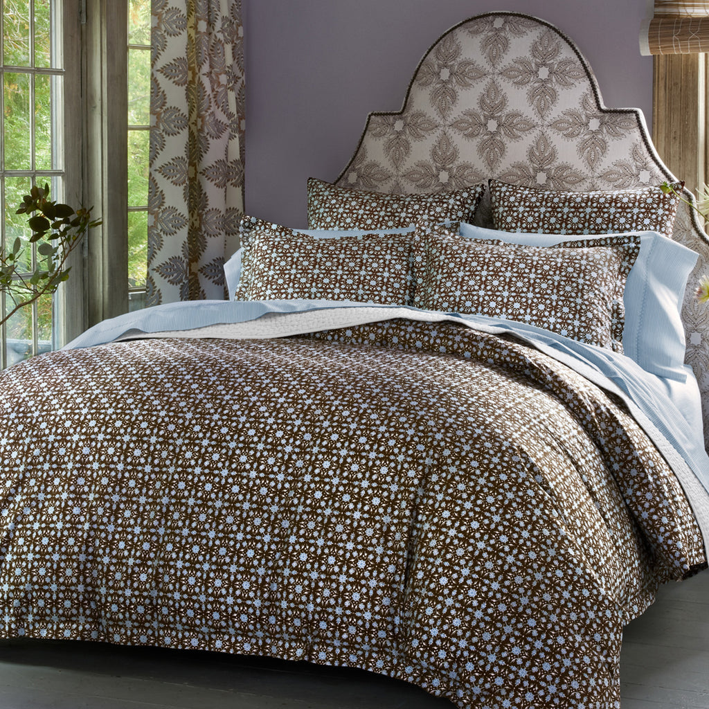 A bedroom showcasing the John Robshaw Kaarik Coffee Duvet set, with a patterned headboard and brown and white floral design, is bathed in natural light from a nearby window. A potted plant sits beside the bed, which features an organic cotton duvet insert for extra comfort.
