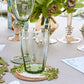 A green-tinted glass tumbler and wine glass sit on a table with Kim Seybert Oasis Coasters Set, floral arrangements, and a printed menu.