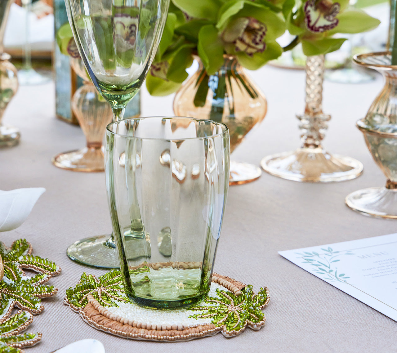 A green-tinted glass tumbler and wine glass sit on a table with Kim Seybert Oasis Coasters Set, floral arrangements, and a printed menu.
