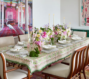 A dining table set with floral arrangements, candles, and tableware. The vibrant hand-painted Kim Seybert Mira Tablecloth in green and pink hues adds a touch of elegance, while a small decorative green rabbit sits at the center, perfect for garden parties.