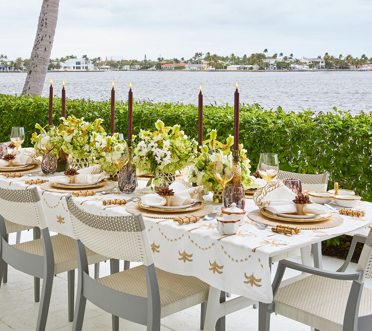 An Embroidered Palm Tablecloth by Kim Seybert dresses the outdoor dining table, paired with floral centerpieces, gold accents, folded napkins, and tall dark candles, set by a waterfront with house and greenery views.