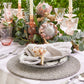 Elevate outdoor entertaining with a handcrafted Kim Seybert Driftwood Gray Placemat, white plate, folded napkin, seashell napkin ring, pink glassware, candles, and a floral centerpiece featuring artichokes and greenery.