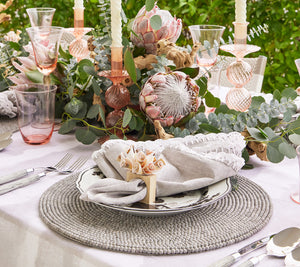 A decorated dining table features a floral centerpiece, candles, intricate linens, and elegantly arranged place settings on Kim Seybert Driftwood Placemats in Gray, set against a lush green backdrop.