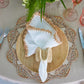 A bohemian table setting features the Kim Seybert Shell Edge Napkin Set, a woven raffia-border placemat, wooden plate, wicker napkin ring, bamboo-handle cutlery, pink glassware, and a beige tablecloth.