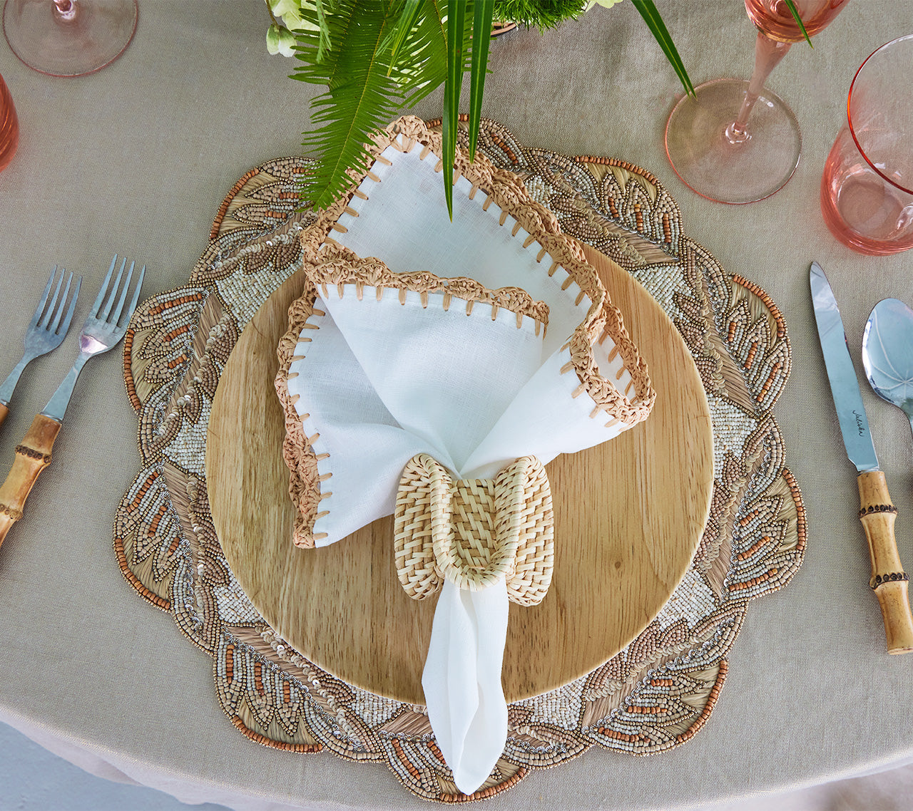 A bohemian table setting features the Kim Seybert Shell Edge Napkin Set, a woven raffia-border placemat, wooden plate, wicker napkin ring, bamboo-handle cutlery, pink glassware, and a beige tablecloth.
