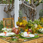 A tropical table setting features bamboo chairs, green plates on a Kim Seybert Tropicana Placemat, napkins, decorative glasses, and a centerpiece of pineapples, bananas, and yellow flowers.