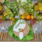 Create a tropical table setting with Kim Seybert’s Tropicana Placemat, a wooden plate, napkin, and a centerpiece featuring bananas, green orchids, and assorted fruits.