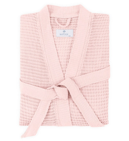 The Kiran Robe by Matouk, shown folded with a matching belt on a white background, is crafted from luxurious light pink Turkish waffle fabric and features the Matouk label on the collar.