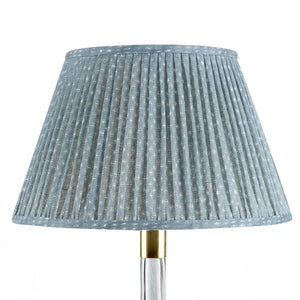 The John Rosselli Fermoie Blue Figured Lampshade features a pleated blue patterned fabric, paired with a lamp that has a clear base and brass neck.