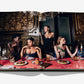Four women in elegant dresses are gathered around a lavishly adorned dining table at Laperouse, the legendary Parisian restaurant celebrated for its sumptuous interiors. Ornate walls, graceful candles, and delectable desserts enhance the scene featured in this open book photo spread from Laperouse: A Parisian Icon Since 1766 by Assouline, capturing the enduring charm of this historic Paris wine cellar.