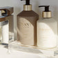 Two L’Avant Collective Ambre Santal Collection bottles—hand soap and hand lotion—rest on a clear tray by the sink, their sandalwood scent mingling with soft sunlight and shadows.