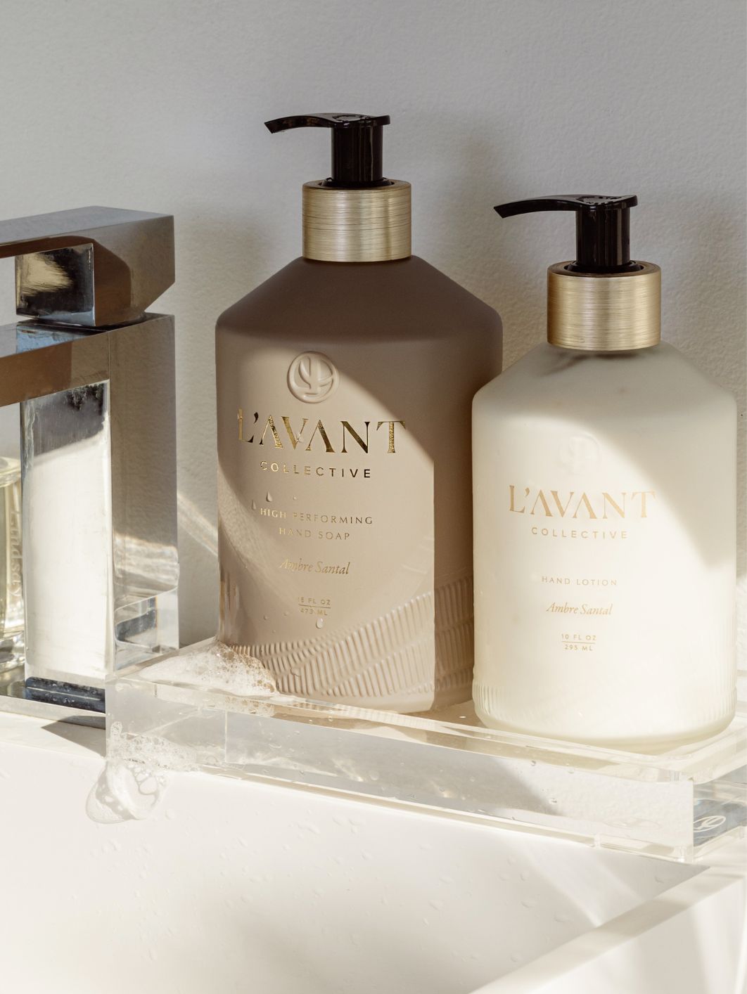 Two L’Avant Collective Ambre Santal Collection bottles—hand soap and hand lotion—rest on a clear tray by the sink, their sandalwood scent mingling with soft sunlight and shadows.