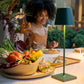 A bowl of fresh vegetables and a Zafferano America Poldina Pro Lamp are on the table; a woman dines and smiles in the background, showcasing the lamp’s portable convenience for indoor or outdoor use.