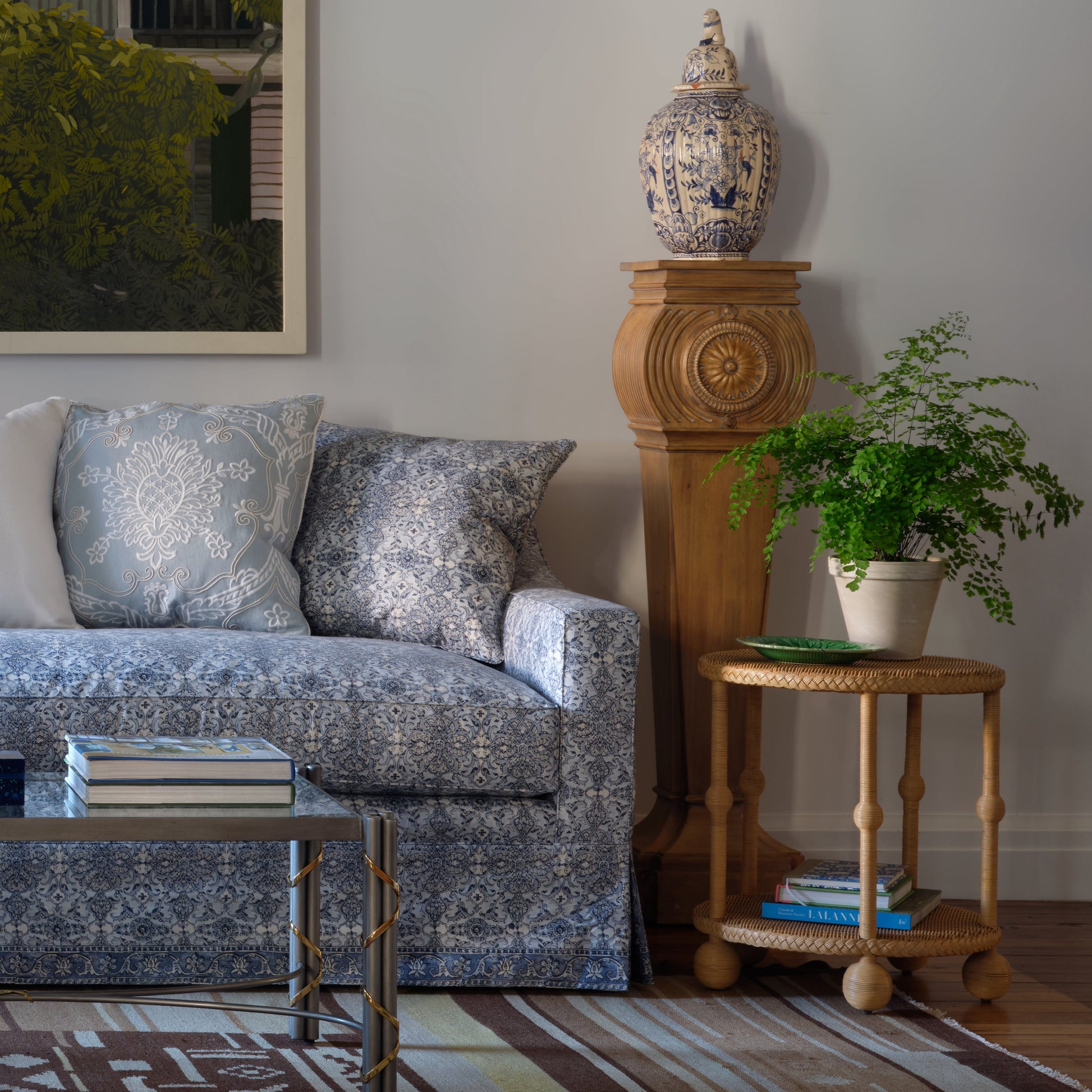 A blue-patterned sofa with matching pillows sits next to the Bunny Williams Lucas Drinks Table, which holds a potted fern; behind them stands a wooden pedestal with a blue-and-white vase.