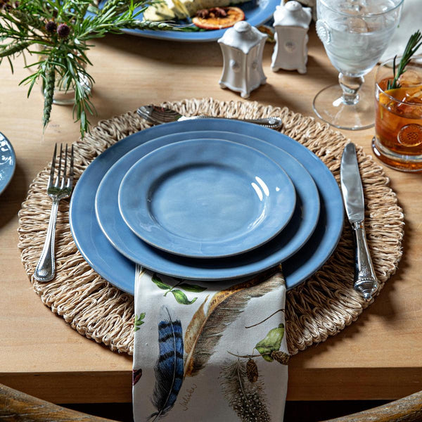 A wooden table set with blue ceramic plates, silver cutlery, a bird-patterned napkin, drinks in glasses, and a Juliska Straw Loop Placemat Collection adding natural texture.